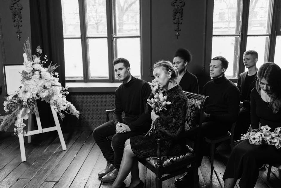 Inside a dimly lit room with large windows, a group of six people sits silently in chairs, dressed in dark clothing suitable for a somber occasion. In the foreground, a woman with short hair, wearing 