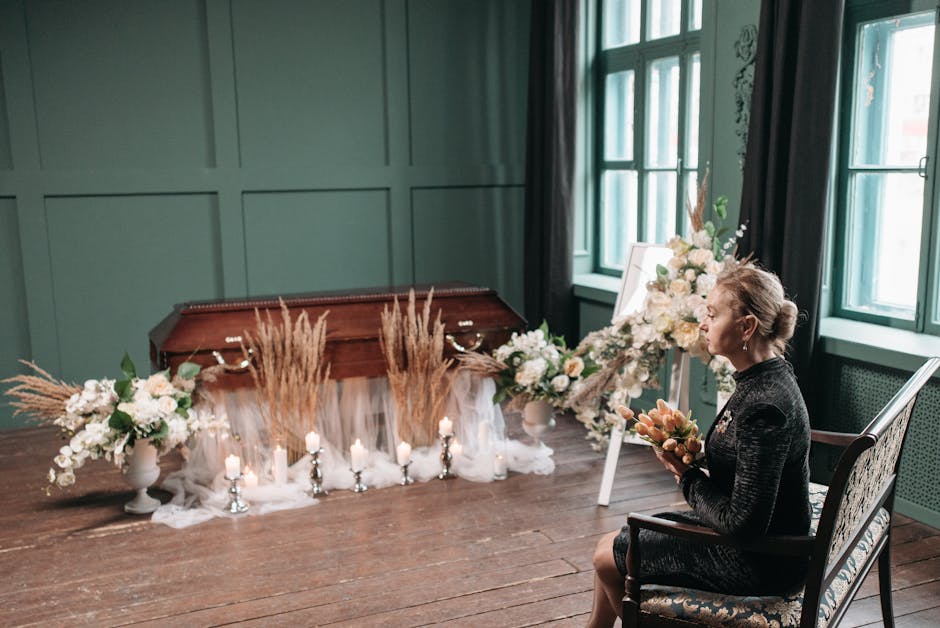 A woman seated indoors on a vintage armchair, holding a floral bouquet featuring soft peach, blush pink, and ivory flowers, in front of a sympathetic floral arrangement on an easel. The arrangement in