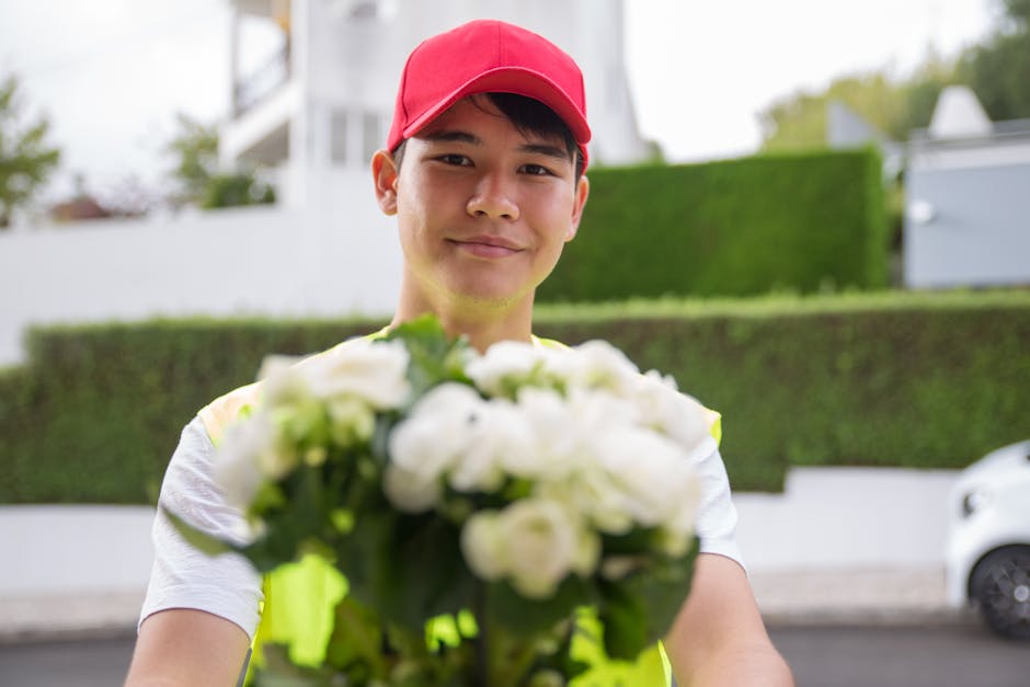 A delivery person wearing a red cap and a neon yellow vest is smiling softly while holding a bouquet of fresh white flowers, including roses and ranunculus, wrapped in clear cellophane. The bouquet is