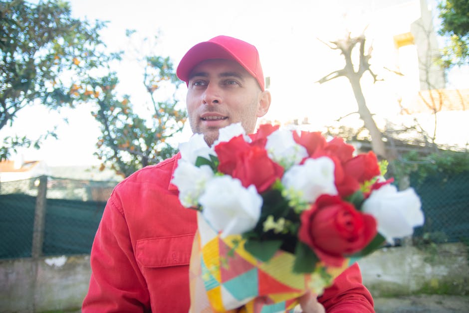 A male florist dressed in a red uniform and matching cap holding a colorful bouquet composed of red and white roses, set outdoors during daytime. The bouquet features a mix of fresh roses and greenery