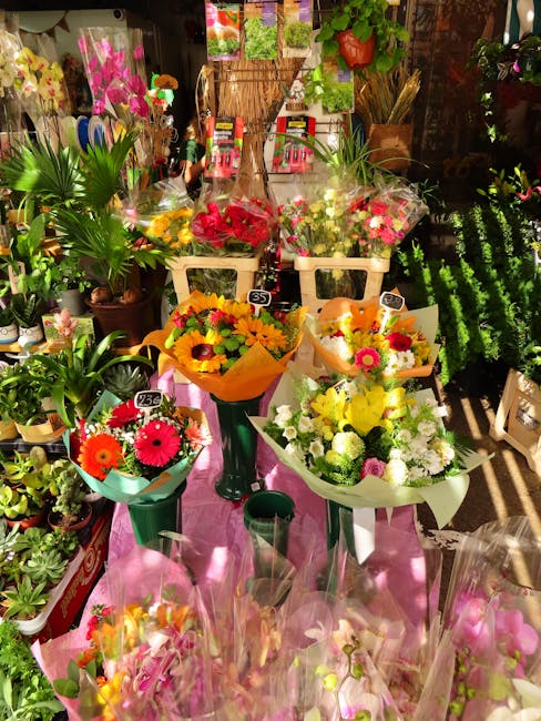 An indoor display of fresh flower bouquets arranged in various vases and wrapping, featuring bright yellow, orange, pink, red, and white blooms including gerbera daisies, chrysanthemums, and roses. Th