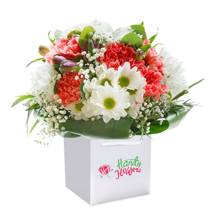 Standing upright within a crisp white gift bag, this bouquet is a vivid showcase of florist expertise, reminiscent of Hanwell's friendly charm and lush green spaces like Elthorne Park. Lush coral and peach carnations, celebrated for their abundant, gently ruffled petals, steal the scene with their velvety textures and vibrant shades of red, pink, and orange. These blooms are thoughtfully paired with pure white daisies, their yellow-lime eyes twinkling with optimism, adding cheerful contrast that conjures scenes of open local meadows. Interwoven throughout the arrangement, small sprays of baby's breath appear as airy, cloud-like accents, bringing softness and subtle movement to the composition. On the bouquet's left side, a few buds tinged with purple lay nestled between the blooms, hinting at fresh beginnings and adding depth to the overall shape. Framing this floral ensemble, robust, deep green leaves spread gracefully, serving as a natural collar that enhances the sense of fullness and structure, much like the community spirit of Hanwell's neighborhoods. The bouquet is shaped to perfection-rounded, abundant, and harmonious-inviting both admiration and celebration. Sturdy white ribbon handles on the bag ensure effortless transport, whether to a birthday lunch near Church Road or a heartfelt thank-you for a neighbour in Hanwell. Set before a spotless white backdrop, the photograph spotlights each detail with clarity, from texture to colour, communicating the freshness and special intent behind every selection. Ideal for moments of joy, remembrance, or everyday gratitude, this arrangement symbolises both Hanwell's natural beauty and the attentive hand of its local florists.