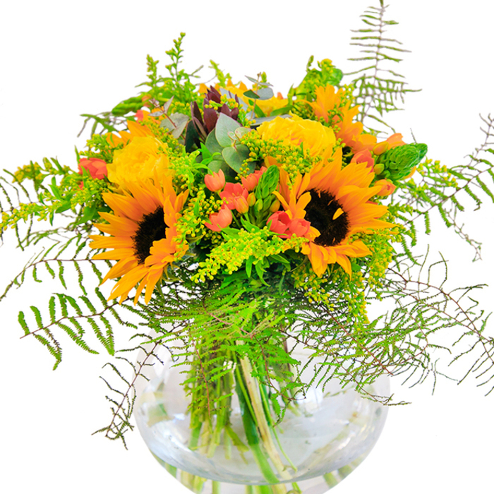 The first impression of this arrangement is a radiant surge of happiness-sunflowers with their dark centers and luminous yellow petals beam beside nestling yellow garden roses, each petal meticulously layered in creamy, spiraled whorls. The bouquet conjures the spirit of summer days in Hanwell, when gardens spill over with colour and life. Lush, feathery fern fronds and airy tendrils of filler-suggestive of goldenrod's tiny yellow clusters-arc outward, softening the boundaries and inviting the eye to wander. Clusters of glossy scarlet Hypericum berries glitter between the blooms, their roundness a vivid counterpoint to the softness of the petals. Fragrant eucalyptus leaves, with their cool blue-green tones, offer subtle highlights and a rustic scent, hinting at the freshness of nearby Osterley Park woods. The fishbowl-like glass vase, clear and modern, puts each green stem on display, adding an extra touch of brightness as they shimmer in water. This bouquet is a tribute to Hanwell's warm-hearted community-a piece to mark joyful milestones such as birthdays, anniversaries, or spontaneous celebrations, infusing the home with light and seasonal beauty.