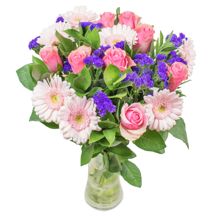 An eye-catching, artistically arranged bouquet is displayed in a clear glass vase, photographed against a clean white backdrop to emphasise each detail. Approximately seven to nine pink roses range from tightly closed buds to newly opened blooms, their classic spiral forms recalling the timeless romance felt along Hanwell's canals. Interwoven are five to seven pale pink gerbera daisies, each with a large, dainty face and radiating petals blending towards barely-there white centres, their freshness conjuring local springtime mornings. Clusters of vivid purple statice nestle among the roses and gerberas, providing striking visual interest and echoing the wildflowers often seen near the River Brent. Lush, deep green foliage surrounds the blossoms, creating abundant volume and a sense of vitality similar to Hanwell's peaceful green spaces. The arrangement's stems and water are fully visible through the transparent vase, emphasizing the freshness and natural beauty of the flowers. Thoughtfully designed for occasions such as birthdays, anniversaries, Mother's Day, or simply to bring cheerful warmth into a room, this bouquet from Flowers Hanwell conveys joy, celebration, and heartfelt sentiment in every exquisite detail.